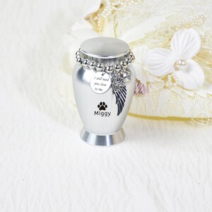 May include: A silver pet urn with a beaded bracelet around the top. The urn has a paw print and the name "Miggy" printed on it. A charm with a wing and the words "I still need you close to me" is attached. The urn is a memorial item.