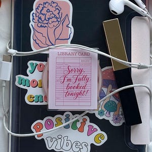 May include: A black tablet case covered in various stickers, including one that reads "Sorry, I'm fully booked tonight!" and another saying "positive vibes." A pair of peach-tinted sunglasses, white wired earbuds, a gold-toned pen, and a bright pink book-shaped case.