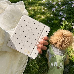 Könnte beinhalten: Eine weiße Tablet-Hülle mit schwarzem Polka-Dot-Muster und goldenen Initialen. Die Hülle wird über einer grünen, grasbewachsenen Fläche mit kleinen weißen Blumen gehalten. Im Hintergrund ist ein Hund mit grünen Latzhosen mit dem Text "AND" zu sehen.