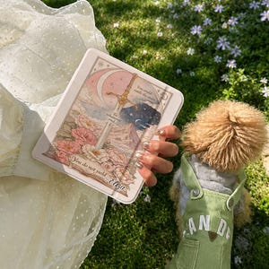 May include: A rectangular card with an illustration of a sword, moon, and flowers. The card has text that reads "You do not yield Eliza" and "You could rattle the stars". A dog in green overalls is in the background.