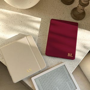 Può includere: Un e-reader bianco che mostra del testo, accompagnato da una custodia beige chiusa con le iniziali dorate AS e una custodia magenta scuro chiusa con le iniziali dorate BL. I dispositivi sono rettangolari con angoli arrotondati e finitura opaca.