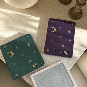Pode incluir: Duas capas para leitor de e-books em verde-azulado e roxo, ambas com padrões celestiais de luas e estrelas em folha de ouro. A capa verde-azulado tem as iniciais AS, enquanto a roxa exibe as iniciais BL. Um leitor de e-books branco é visível.