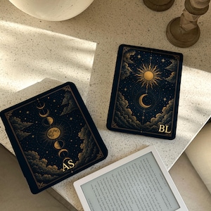Pode incluir: Duas capas pretas para leitor de e-books com ilustrações celestiais douradas de sóis, luas e nuvens. Uma capa exibe as letras AS, a outra BL. Um leitor de e-books branco com texto visível está posicionado abaixo das duas capas.