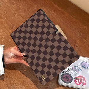 Hand holding a chocolate checkerboard Kindle case with personalised gold initials, shown from above on a wooden surface