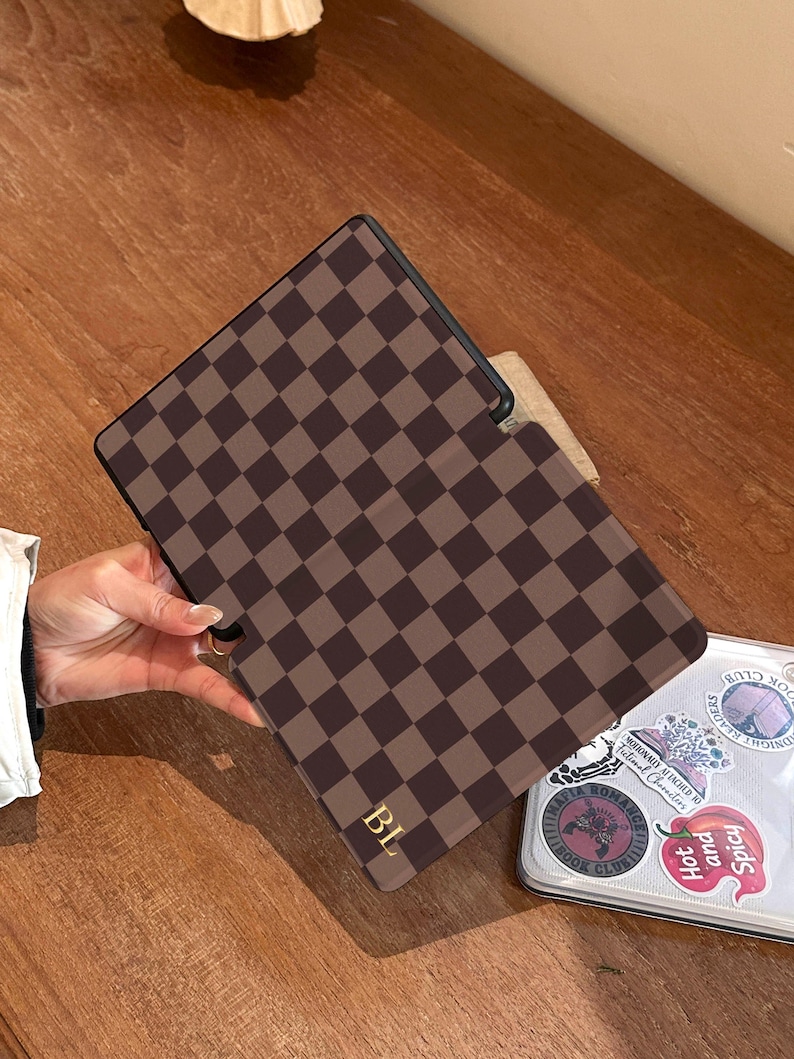 Hand holding a chocolate checkerboard Kindle case with personalised gold initials, shown from above on a wooden surface