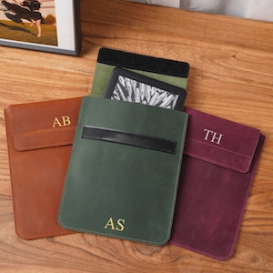 Puede incluir: Tres fundas rectangulares para lectores electrónicos en color tostado, verde oscuro y morado intenso. Cada funda cuenta con un cierre de solapa con una correa horizontal e iniciales grabadas en oro: AB en la tostada, AS en la verde y TH en la morada.