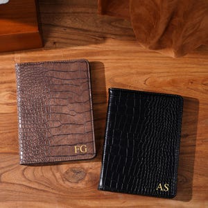 May include: Two passport holders with a crocodile skin texture. One is brown with gold initials "FG", the other is black with gold initials "AS". The holders are displayed on a wooden surface.