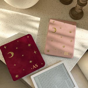 May include: Two e-reader covers in deep red and light pink, both featuring gold foil celestial motifs of a crescent moon and stars. The red cover is personalized with the letters AS, while the pink cover features the letters BL in gold foil.