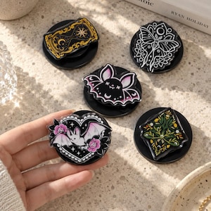 May include: Five circular phone grips featuring acrylic tops with various designs: a gold and black hourglass, a white moth, a black bat, a heart-shaped bat with pink flowers, and a green diamond-shaped botanical pattern with small yellow flowers.
