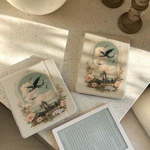 May include: Three cream-coloured e-reader cases with a fantasy design featuring a dragon, castle, and floral accents. A white e-reader displaying text is also present. The cases have the name "Elsa" printed on them.