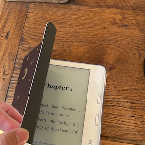 May include: A white Kobo e-reader displaying Chapter 1 of the book Court of Thorns and Roses. A dark brown cover with gold moon and star illustrations is partially open, revealing the screen text which reads, The forest had become a path of snow and ice.