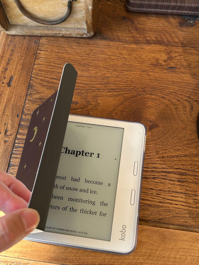 May include: A white Kobo e-reader displaying Chapter 1 of the book Court of Thorns and Roses. A dark brown cover with gold moon and star illustrations is partially open, revealing the screen text which reads, The forest had become a path of snow and ice.