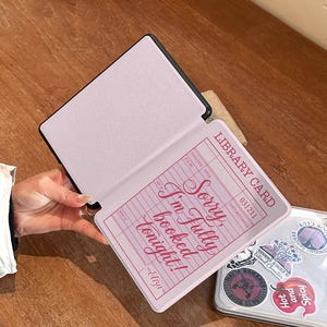 Hand holding an open Kindle case showing the retro library card design printed inside the front cover. Slim protective Kindle cover designed for comfortable reading sessions. Bookish aesthetic Kindle case compatible with Kindle Paperwhite and Kindle 6 devices.