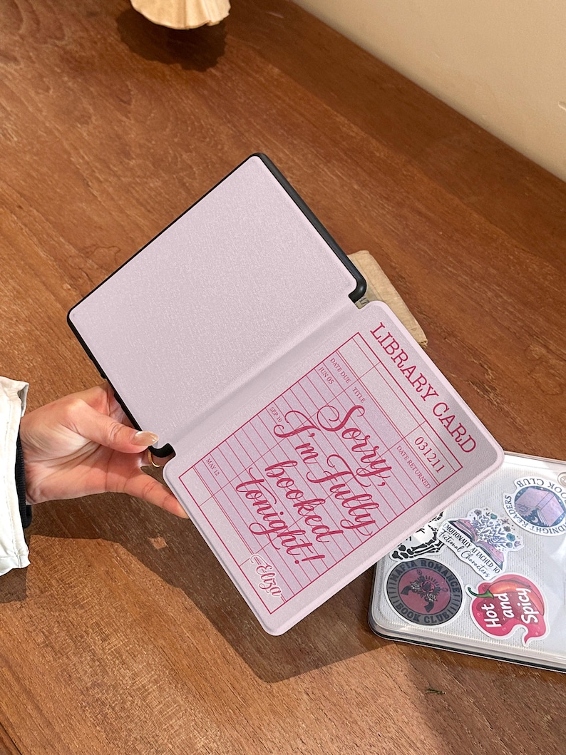 Hand holding an open Kindle case showing the retro library card design printed inside the front cover. Slim protective Kindle cover designed for comfortable reading sessions. Bookish aesthetic Kindle case compatible with Kindle Paperwhite and Kindle 6 devices.