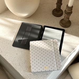 May include: A black e-reader device featuring a protective cover with a white background and a black polka dot pattern. The cover includes a gold monogram with the letters BL at the bottom. The interior of the case is black with a hand strap.