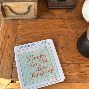 May include: A white and blue library card-themed item with the text "Books Are My Love Language" in a cursive font. The item has a light green background and is on a wooden surface. The item is a decorative piece.