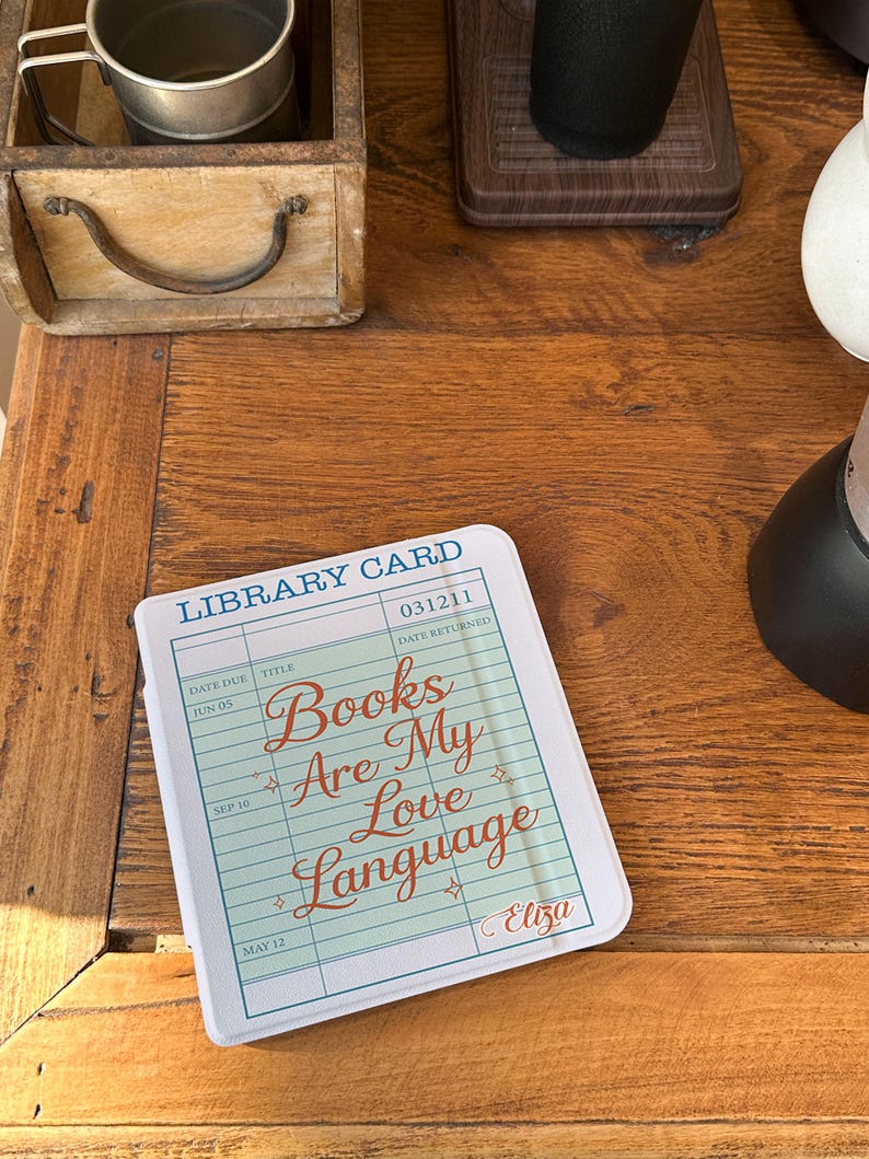 May include: A white and blue library card-themed item with the text "Books Are My Love Language" in a cursive font. The item has a light green background and is on a wooden surface. The item is a decorative piece.