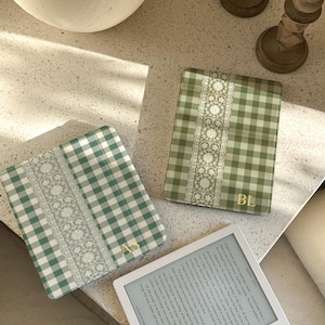 May include: Two e-reader covers featuring a sage green and white gingham pattern with a vertical white lace-style floral strip down the center. One cover displays the gold initials AS, while the other displays the gold initials BL.