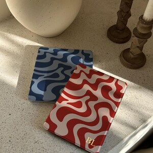 Puede incluir: Dos tarjeteros rectangulares con patrones ondulados. Uno es rojo con remolinos blancos, el otro es azul con remolinos azul oscuro. El tarjetero rojo tiene las iniciales doradas "BL".
