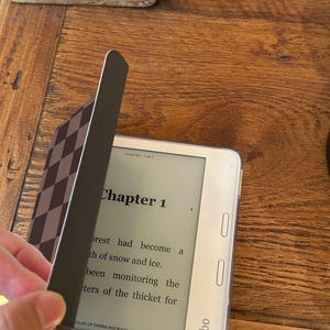 May include: A white Kobo e-reader displaying Chapter 1 of the book A Court of Thorns and Roses. The device is partially covered by a dark brown and light brown checkered magnetic cover that is being lifted to reveal the screen text.