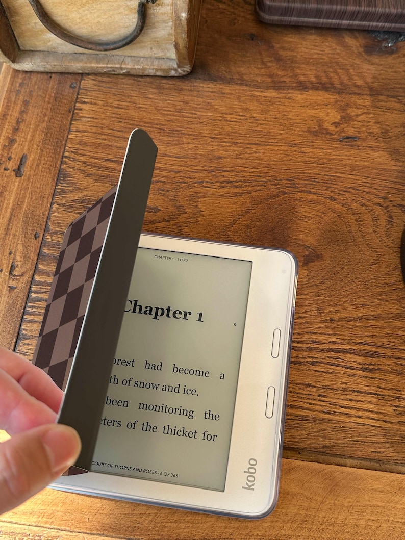 May include: A white Kobo e-reader displaying Chapter 1 of the book A Court of Thorns and Roses. The device is partially covered by a dark brown and light brown checkered magnetic cover that is being lifted to reveal the screen text.
