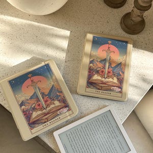 May include: Three cream-colored e-reader cases with a fantasy illustration of a sword, open book, and mountains. The cases have the text "THE READER" and the brand name "Eliza". A white e-reader with text is also visible.