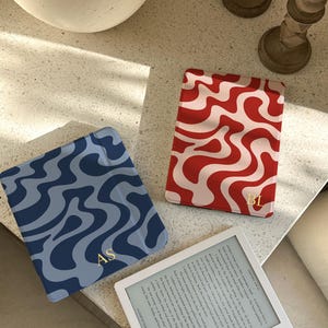 Puede incluir: Tres fundas decorativas para lectores electr&oacute;nicos con dise&ntilde;os ondulados. Una es azul con remolinos azul oscuro e iniciales doradas "AS". Otra es roja y blanca, con iniciales doradas "BL". Tambi&eacute;n se ve un lector electr&oacute;nico blanco.