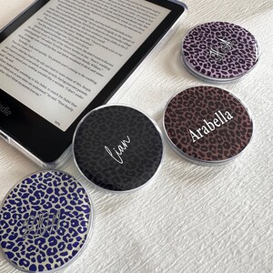 May include: Four circular, flat, clear-rimmed phone grips featuring leopard print patterns in purple, black, brown, and blue. Each grip displays a custom name in white script: Arabella, Lian, and Dion. One grip is placed next to an e-reader device.