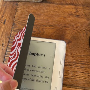 Puede incluir: Un lector electr&oacute;nico Kobo blanco con una funda protectora con un dise&ntilde;o ondulado rojo y blanco. La pantalla muestra el texto del "Cap&iacute;tulo 1" de un libro. La funda se est&aacute; abriendo para revelar la pantalla.