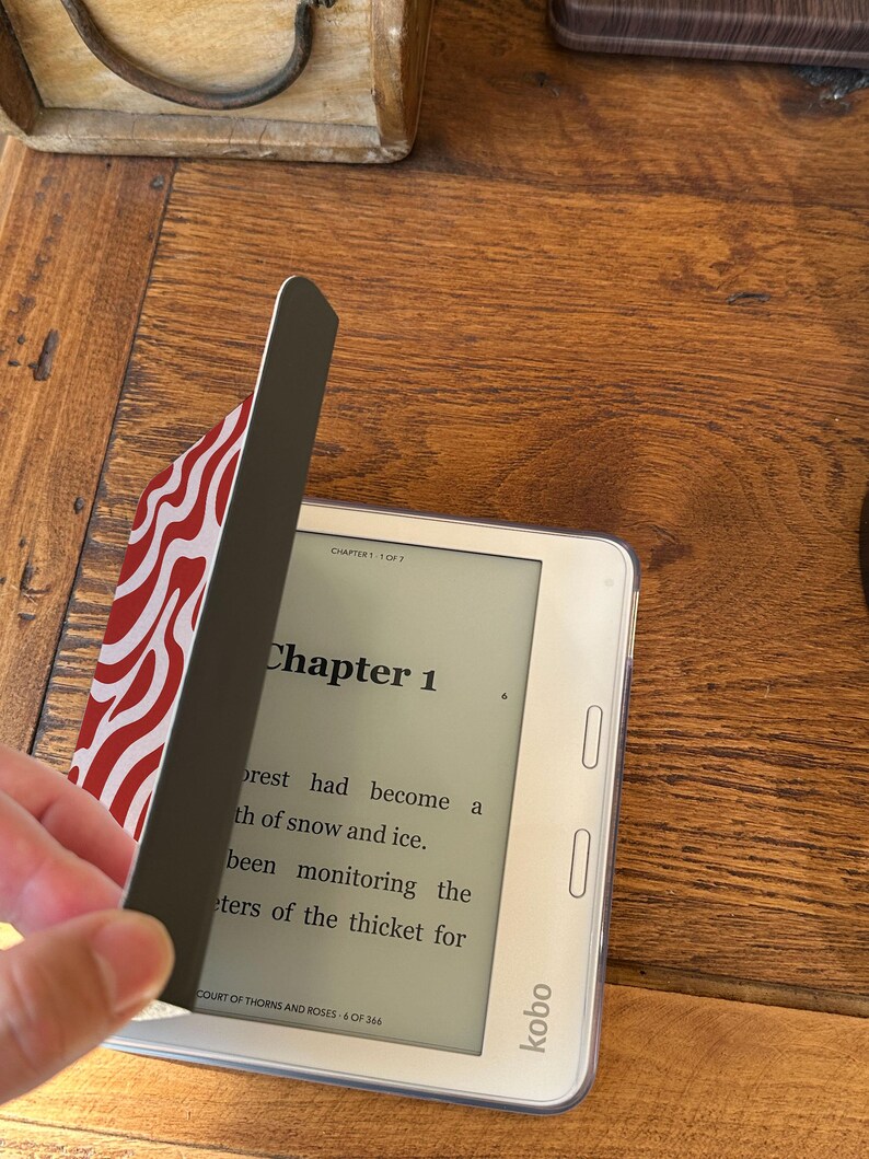 Puede incluir: Un lector electr&oacute;nico Kobo blanco con una funda protectora con un dise&ntilde;o ondulado rojo y blanco. La pantalla muestra el texto del "Cap&iacute;tulo 1" de un libro. La funda se est&aacute; abriendo para revelar la pantalla.