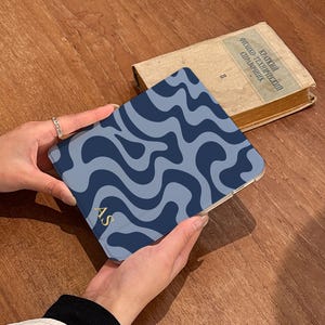 Puede incluir: Una funda con estampado de ondas azules y marino con las iniciales doradas "AS" es sostenida. Un libro antiguo con texto ruso est&aacute; en el fondo. La funda es cuadrada y parece ser una funda para tableta o tel&eacute;fono.