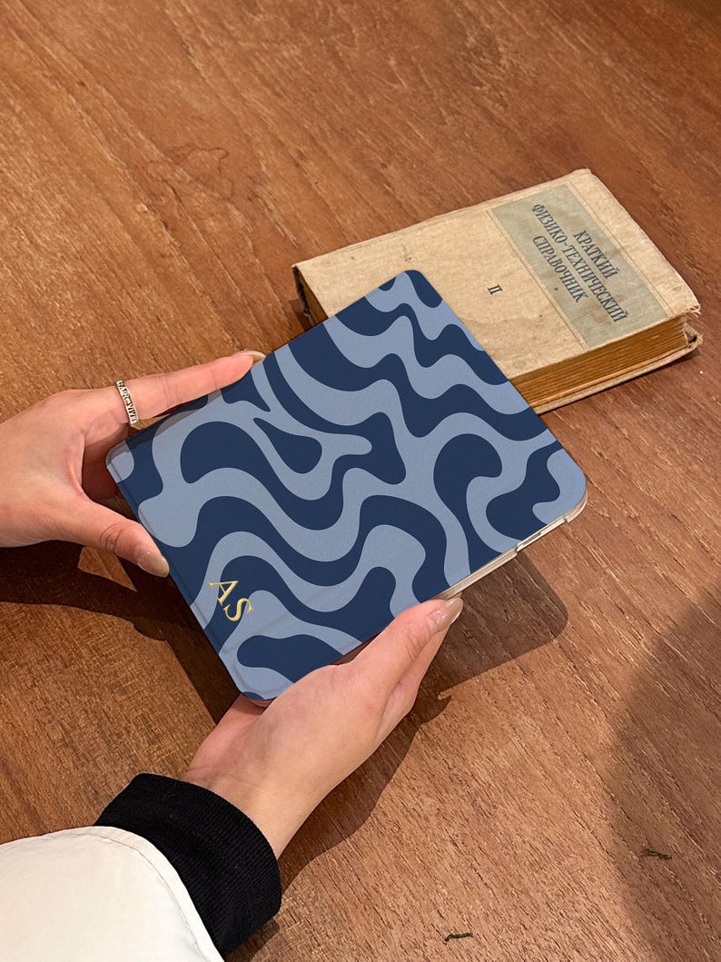 Puede incluir: Una funda con estampado de ondas azules y marino con las iniciales doradas "AS" es sostenida. Un libro antiguo con texto ruso est&aacute; en el fondo. La funda es cuadrada y parece ser una funda para tableta o tel&eacute;fono.