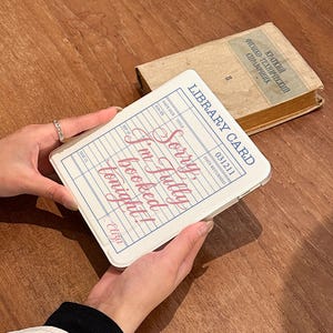 May include: A white phone case designed as a library card, featuring the text "Sorry I'm fully booked tonight!" in pink script. The case has a blue border and a vintage style. An old book is in the background.