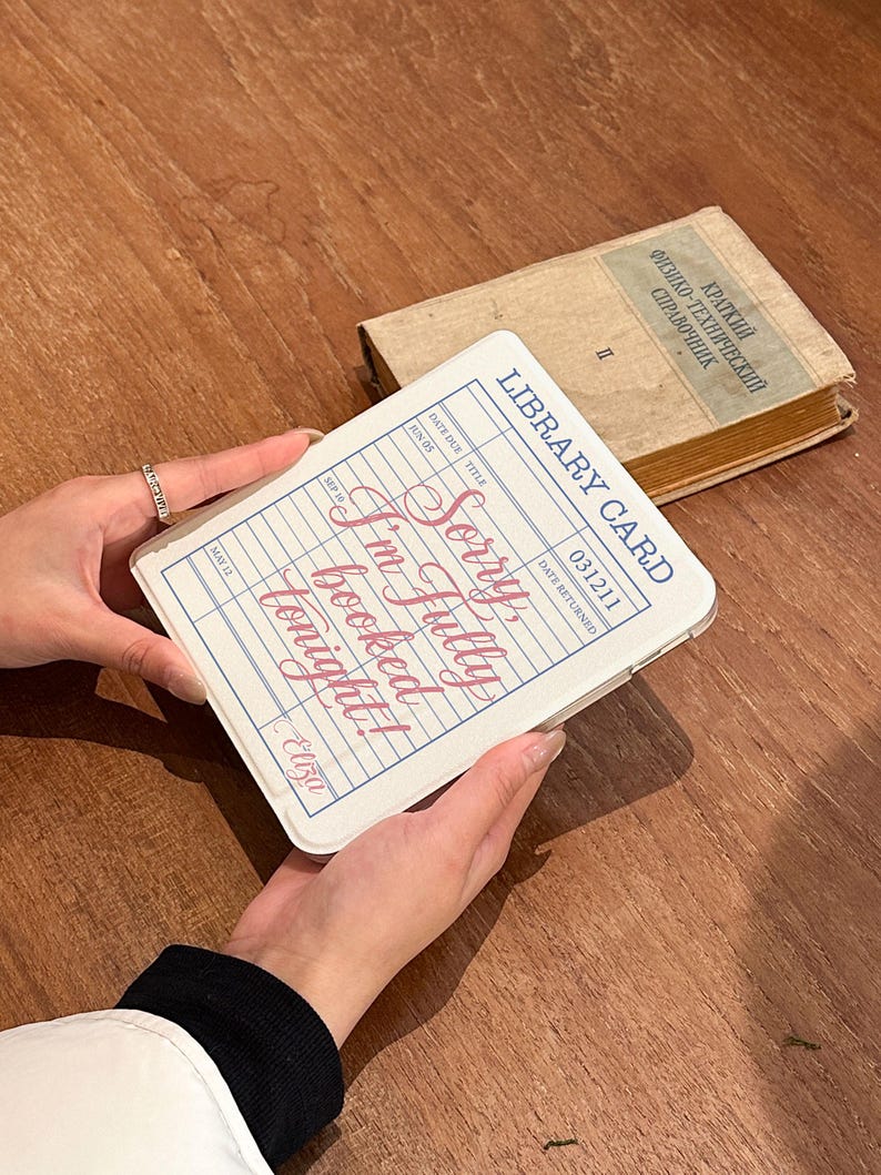 May include: A white phone case designed as a library card, featuring the text "Sorry I'm fully booked tonight!" in pink script. The case has a blue border and a vintage style. An old book is in the background.