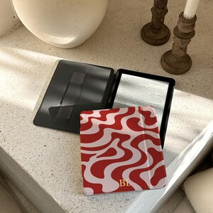 Puede incluir: Un lector electr&oacute;nico negro abierto con una pantalla que muestra texto, junto a una funda con estampado rojo y blanco. La funda tiene un dise&ntilde;o ondulado e iniciales doradas. Tambi&eacute;n se ve una funda negra con una correa de mano.