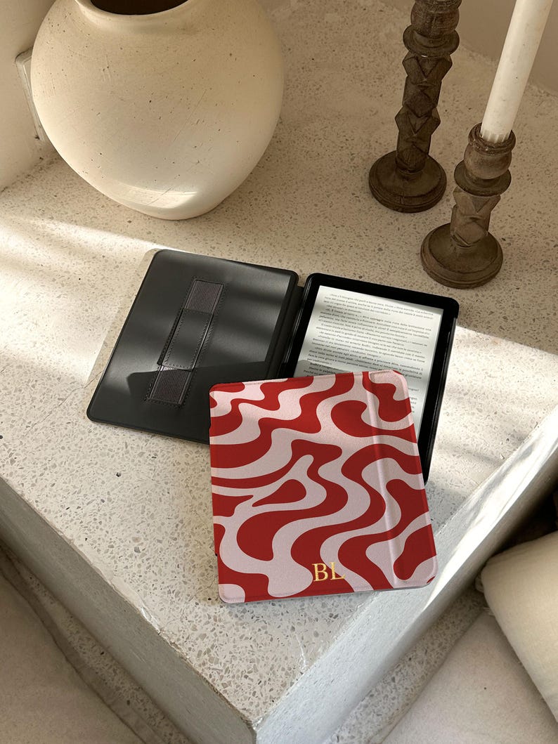 Puede incluir: Un lector electr&oacute;nico negro abierto con una pantalla que muestra texto, junto a una funda con estampado rojo y blanco. La funda tiene un dise&ntilde;o ondulado e iniciales doradas. Tambi&eacute;n se ve una funda negra con una correa de mano.