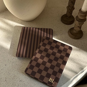 Chocolate checkerboard Kindle case with gold initial detail, placed on a wooden table with glassware in warm sunlight, cosy reading mood