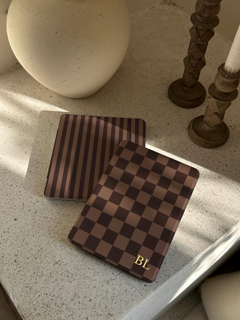 Chocolate checkerboard Kindle case with gold initial detail, placed on a wooden table with glassware in warm sunlight, cosy reading mood