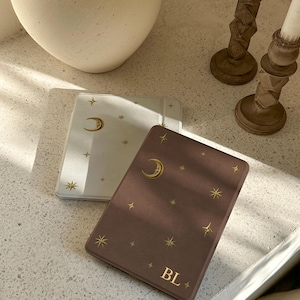 May include: Two rectangular e-reader cases, one white and one dark mauve, both featuring a gold foil celestial pattern of a crescent moon and various stars. The dark mauve case is personalized with the gold foil initials BL at the bottom.