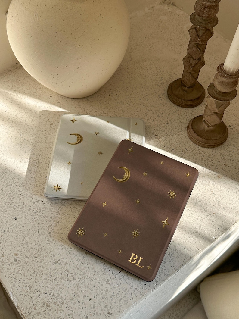 May include: Two rectangular e-reader cases, one white and one dark mauve, both featuring a gold foil celestial pattern of a crescent moon and various stars. The dark mauve case is personalized with the gold foil initials BL at the bottom.