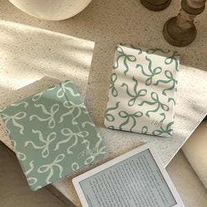 Pode incluir: Três capas de livros com um padrão de laço verde claro e branco, com o nome "Eliza" impresso em duas delas. Um leitor de e-books branco exibe texto. As capas e o leitor estão em uma superfície clara.