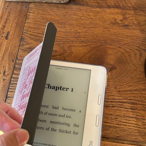 May include: A white Kobo e-reader with a clear protective case, displaying text from "Court of Thorns and Roses." A black magnetic bookmark with "Library Card" text is partially covering the screen. The e-reader is on a wooden surface.
