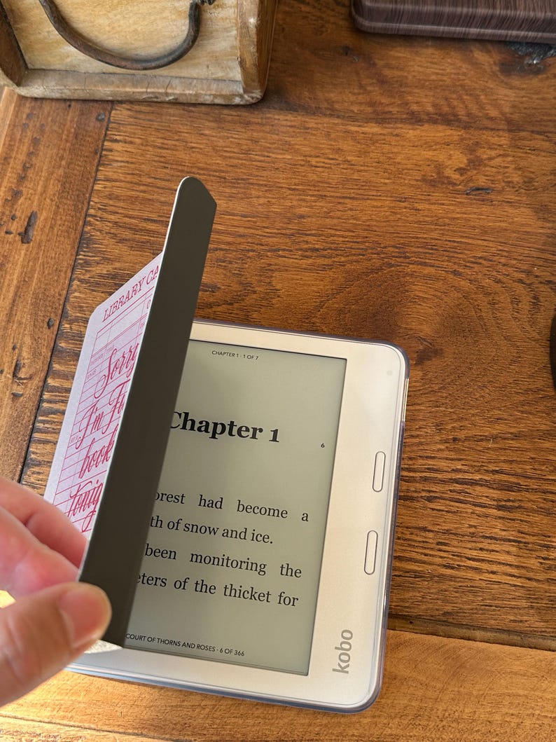 May include: A white Kobo e-reader with a clear protective case, displaying text from "Court of Thorns and Roses." A black magnetic bookmark with "Library Card" text is partially covering the screen. The e-reader is on a wooden surface.