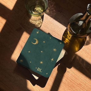 May include: A dark green square notebook with a gold foil crescent moon and star pattern on the cover. The letters BL are printed in gold at the bottom center. The notebook is positioned next to a clear glass and a dark glass bottle.