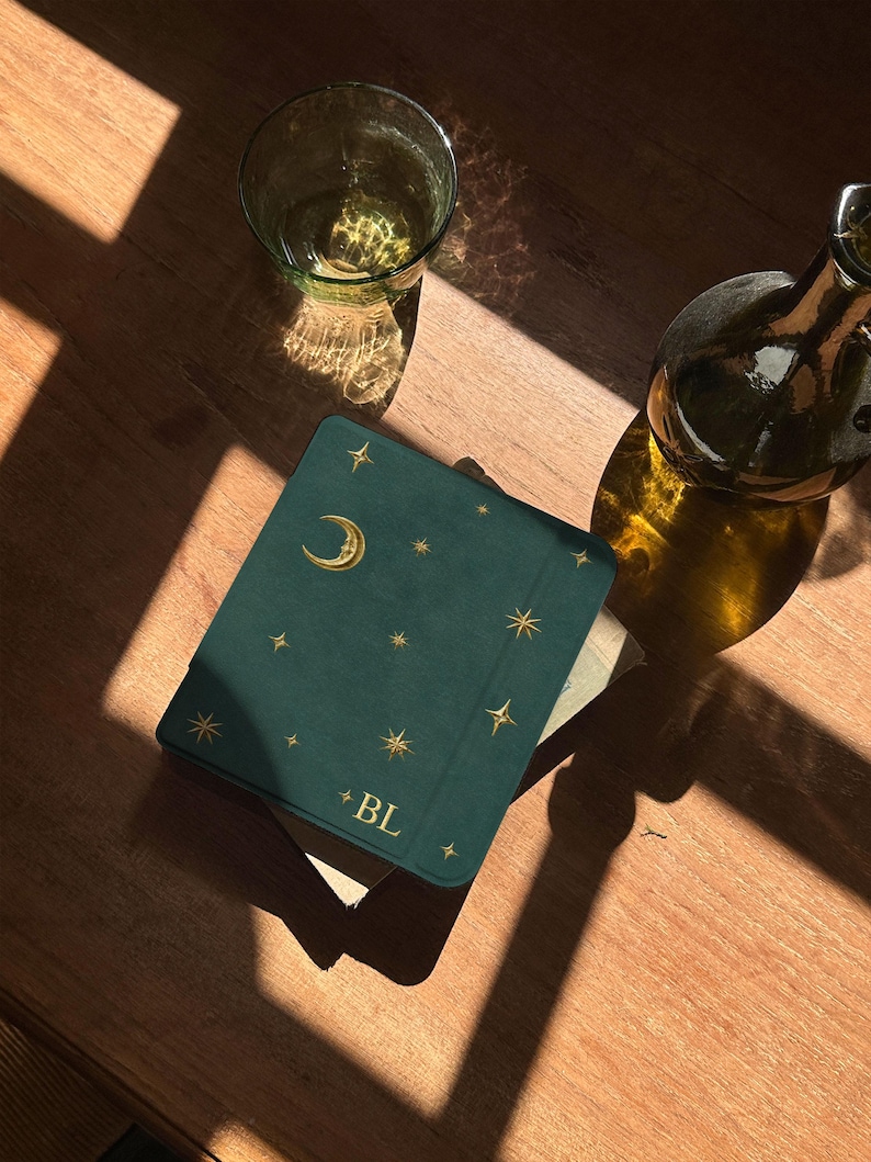May include: A dark green square notebook with a gold foil crescent moon and star pattern on the cover. The letters BL are printed in gold at the bottom center. The notebook is positioned next to a clear glass and a dark glass bottle.