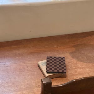 May include: A square coaster featuring a dark brown and black checkerboard pattern resting on top of a small, weathered, light-colored vintage book. The book and coaster are positioned on a smooth, polished wooden surface with a visible grain.