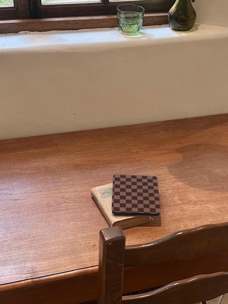 May include: A square coaster featuring a dark brown and black checkerboard pattern resting on top of a small, weathered, light-colored vintage book. The book and coaster are positioned on a smooth, polished wooden surface with a visible grain.