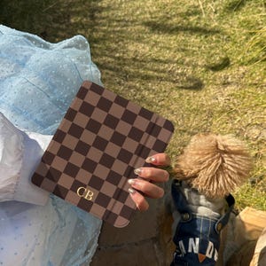 May include: A square electronic reader case featuring a dark brown and light brown checkered pattern with the gold-colored initials CB printed in the bottom left corner. The case has a smooth, matte finish and a slim, rectangular profile.
