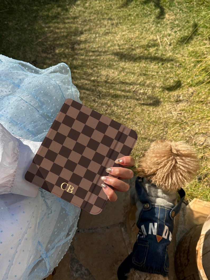 May include: A square electronic reader case featuring a dark brown and light brown checkered pattern with the gold-colored initials CB printed in the bottom left corner. The case has a smooth, matte finish and a slim, rectangular profile.