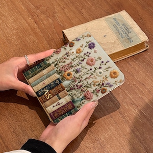 May include: A square phone case with a design of stacked books and a floral arrangement. The case has the initials "AS" in gold. An old book with Russian text is in the background.